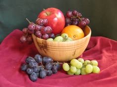 a wooden bowl filled with lots of fruit on top of a red cloth next to grapes and an apple