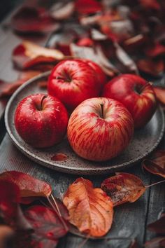 three red apples on a plate surrounded by autumn leaves and fallen leaves, sitting on a wooden table