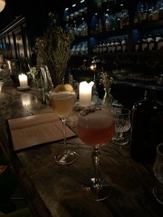 two cocktails are sitting on the bar with candles and books in front of them