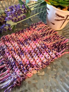 a crocheted dishcloth next to a potted plant on a table with purple flowers