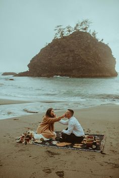 a man kneeling down next to a woman on top of a blanket at the beach