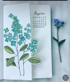 an open notebook with blue flowers on it next to a flower plant and a marker