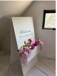 a welcome sign with pink and orange flowers in it sitting on a white sheeted bed