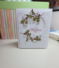 a christmas card with holly leaves and red berries on it, sitting on a desk