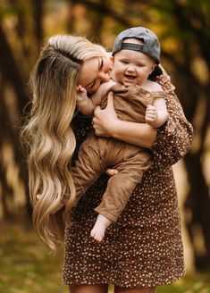 a woman holding a baby in her arms and kissing the child's cheek with trees in the background