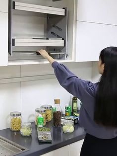 a woman is reaching for something in the kitchen cabinet above the counter top, with spices and condiments around her