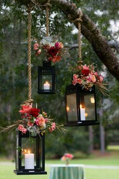 three hanging lanterns with flowers and candles are hung from a tree in front of a table