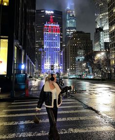 a woman walking across a cross walk in front of tall buildings with lights on at night