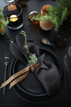 a table set with black plates and napkins, silverware, candles and greenery