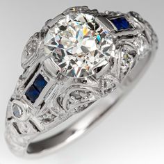 This gorgeous circa 1930s Art Deco engagement ring features a pierced and engraved design and milgrain details. The ring is centered with a 1.30 carat round transitional brilliant cut diamond, in a four-prong setting. The diamond is GIA certified M color and VS1 clarity. The top of the ring is bead set with 16 round single cut diamonds. The shoulders are each channel set with two square step cut lab created sapphires. The ring measures 9.9mm at the top and rises 7.6mm above the finger. The ring is currently a size 6 and we offer complimentary resizing to fit. The ring is platinum and the shank was replaced some time ago with sturdy 14k white gold and looks great. Heirloom Milgrain Round Cut Wedding Rings, Formal Round Diamond Ring With Milgrain Detail, Formal Diamond Ring With Milgrain Detailing, Formal Milgrain Diamond Ring, Wedding Diamond Ring With Milgrain, Luxury Round Milgrain Diamond Ring, Luxury Milgrain Round Diamond Ring, Luxury Round Diamond Ring With Milgrain, Classic Round Diamond Ring With Milgrain Detailing