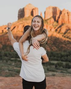 a man holding a woman on his back in front of some mountains and rocks at sunset