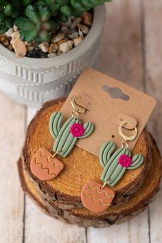 a pair of cactus earrings sitting on top of a piece of wood next to a potted plant