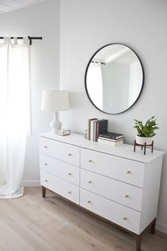 a white dresser with a round mirror on top of it and a lamp next to it