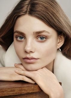 a young woman with blue eyes leaning on a wooden table wearing a white coat and earrings