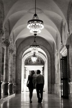 two people are walking down the hallway in an old building with chandeliers hanging from the ceiling