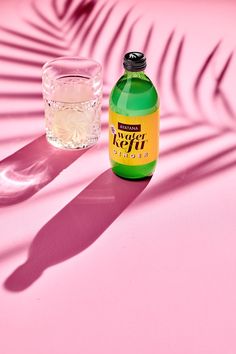 a bottle of alcohol next to a glass on a pink surface with palm leaf shadows