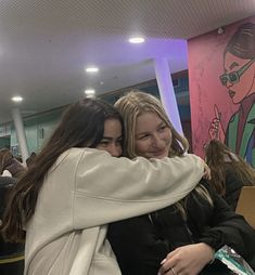 two young women hugging each other in front of a wall with a painting on it