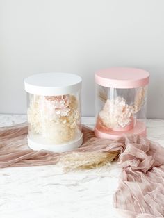 two clear containers with flowers in them sitting on a marble counter top next to a pink and white cloth