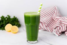 a green smoothie in a tall glass with a striped straw next to lemons and parsley