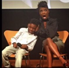 two people sitting on chairs in front of a black wall and one person holding a microphone