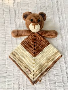 a crocheted teddy bear laying on top of a white bed sheet with a brown and cream blanket