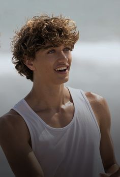 a young man with curly hair and white tank top looking to the side, smiling