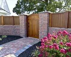 a brick wall with two wooden doors in front of flowers and bushes on the side