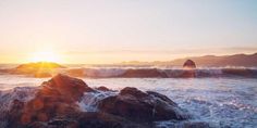 the sun is setting over the ocean with waves crashing on rocks in front of it