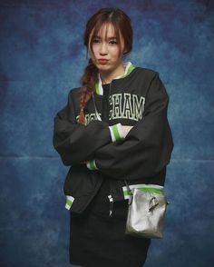 a woman with her arms crossed wearing a black jacket and green striped skirt, standing in front of a blue background