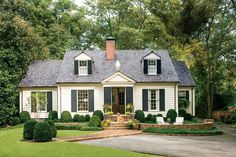 a white house with black shutters in the front yard and landscaping around it, surrounded by trees