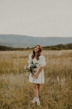 a woman in a white dress holding a bouquet standing in tall grass with mountains in the background