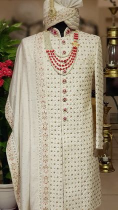 a man's white outfit with red beads on the neck and collar, standing next to a potted plant