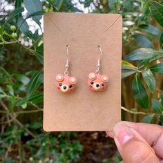 a pair of earrings with pink and orange bear ears on them, sitting in front of a tree