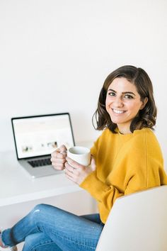 a woman sitting on a chair holding a coffee cup in front of a laptop computer