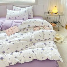 a bed with purple and white comforters in a bedroom next to a lamp on a table