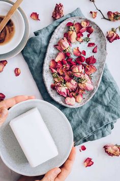 a person holding a soap bar next to a bowl of rose petale and cup of coffee