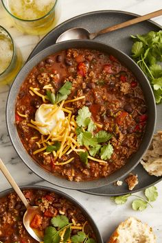 two bowls of chili with sour cream, bread and cilantro on the side