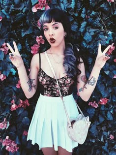 a woman with black hair and tattoos standing in front of flowers holding her hands up