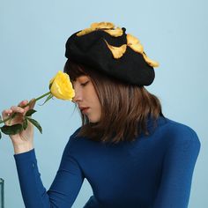 a woman wearing a black hat holding a yellow rose in her right hand and looking down