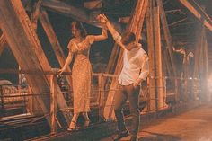 a man and woman dancing on a bridge at night with their hands in the air