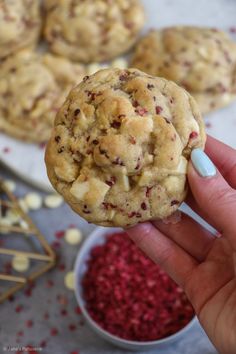 a hand holding up a cookie with crumbles on it next to bowls of red and white sprinkles