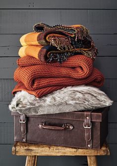 an old suitcase is stacked with blankets and scarves on top of it in front of a gray wall