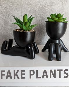 two fake plants in black pots sitting on top of a white shelf next to each other