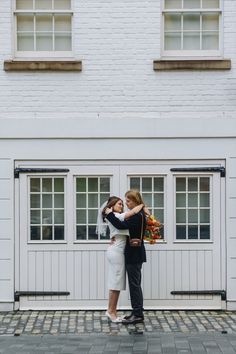 two women embracing each other in front of a white building with windows and brick walkway