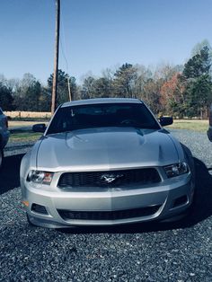 a silver mustang parked in gravel next to other cars and trees on a sunny day