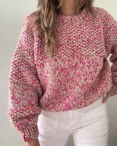 a woman with long hair wearing a pink sweater and white jeans, standing in front of a wall