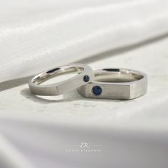 two wedding rings sitting on top of a white cloth covered bed sheet, with one blue diamond in the middle