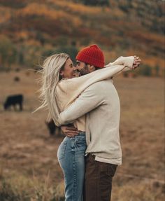 a man and woman embracing in the middle of an open field with cows behind them