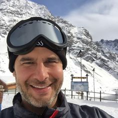 a man wearing ski goggles and smiling at the camera in front of a snowy mountain