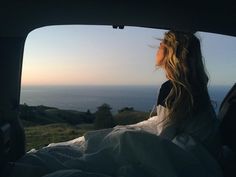 a woman sitting in the back seat of a car looking out at the ocean and hills